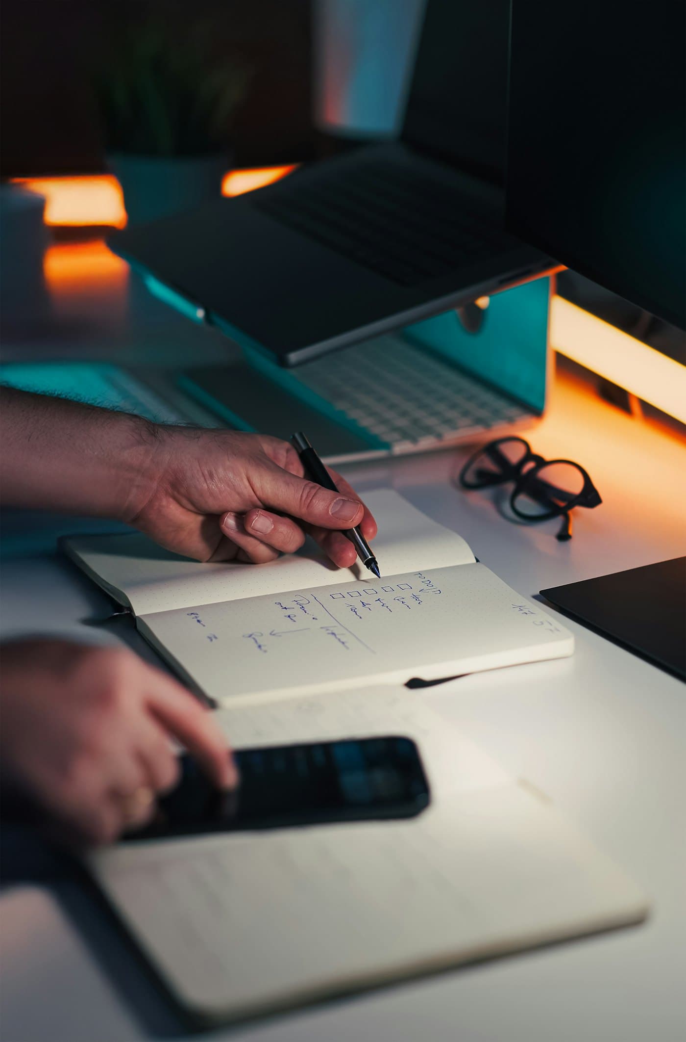 Une main qui note des chiffres dans un carnet, devant un ordinateur portable et un téléphone
